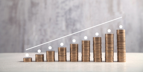 Des piles de pièces monnaie alignée de la plus petite pile à la plus grande, une main dépose une pièce sur la plus grande pile. Un trait courbé suit la pente des piles de monnaie.
